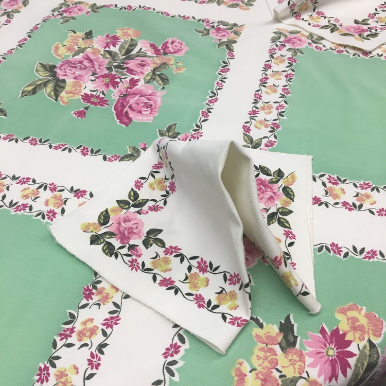 Op de afbeelding: Een wit tafelkleed met een groen bloemenpatroon. Het tafelkleed heeft een rand van roze en gele bloemen. Het tafelkleed is dubbelgevouwen en het bloemenpatroon is aan beide kanten zichtbaar.