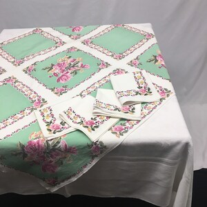 Op de afbeelding: Een wit tafelkleed met een groen en roze bloemenpatroon. Het tafelkleed heeft een ruitpatroon met een bloemendesign in elke ruit. Er zijn vier bijpassende servetten met hetzelfde bloemenpatroon.