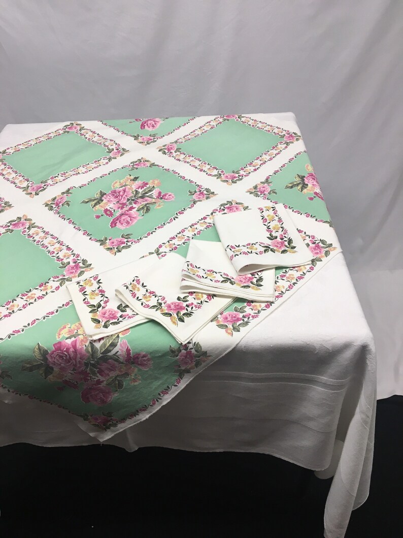 Op de afbeelding: Een wit tafelkleed met een groen en roze bloemenpatroon. Het tafelkleed heeft een ruitpatroon met een bloemendesign in elke ruit. Er zijn vier bijpassende servetten met hetzelfde bloemenpatroon.