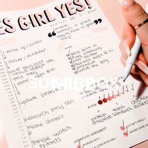 May include: A pink and white planner with the words "Yes Girl Yes" written in black marker. The planner has a daily schedule with tasks and a to-do list. The planner also has a section for end of day assessment with checkboxes for "worked out?" and "got sh't done?" and a space for "cups of water?"