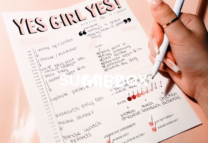May include: A pink and white planner with the words "Yes Girl Yes" written in black marker. The planner has a daily schedule with tasks and a to-do list. The planner also has a section for end of day assessment with checkboxes for "worked out?" and "got sh't done?" and a space for "cups of water?"