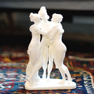 May include: A white sculpture of three figures in an embrace. The figures are in flowing robes and stand on a rectangular base. The sculpture is placed on a patterned rug.