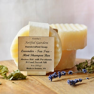 May include: A round bar of handmade soap with a brown label that reads "Keitha's Artful Garden Handcrafted Soap Lavender - Tea Tree - Mint Shampoo Bar Moisture Rich with Pro-vitamin B-5 and Tussah Silk 4 oz. (113 g) Made in Sedalia, VA". The soap is sitting on a wooden surface with dried lavender and mint leaves.