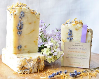 May include: Two bars of handmade soap with lavender and calendula petals. The soap is labeled "Artful Garden Lavender-Lilacs Calendula Soap" and says "skin-loving oils & butters, relaxing scents and flowers".