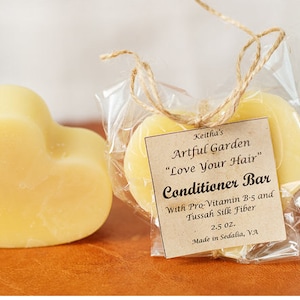 May include: Two heart-shaped bars of conditioner packaged in clear plastic with a brown paper tag. The tag reads "Keitha's Artful Garden "Love Your Hair" Conditioner Bar With Pro-Vitamin B-5 and Tussah Silk Fiber 2.5 oz. Made in Sedalia, VA."