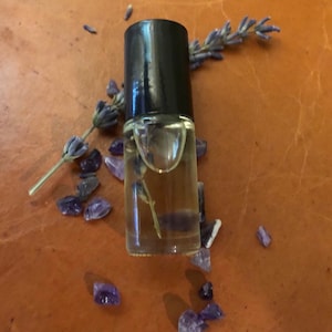 Puede incluir: Una pequeña botella de vidrio transparente con tapa negra llena de un líquido amarillo claro y una ramita de lavanda seca. La botella está rodeada de pequeños cristales de amatista púrpura sobre una superficie de cuero marrón.