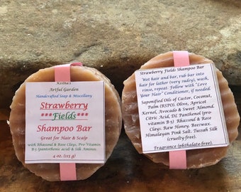 May include: Two round bars of handmade soap, one labeled "Strawberry Fields Shampoo Bar" and the other "Strawberry Fields Shampoo Bar". The soap is brown and has a textured surface. The soap is wrapped in pink and white paper.