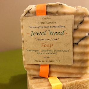 May include: Two bars of handmade soap, one on top of the other. The top bar is wrapped in orange ribbon and has a label that reads "Keitha's Artful Garden Handmade Soap & Miscellany ~Jewel Weed~ "Poison Ivy / Oak" Soap Wild-crafted Jewelweed, French Green Clay, Essential Oils 4 oz. Made in Sedalia, VA".