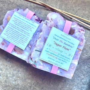 May include: Two handcrafted soaps, labeled "Over The Rainbow" and "Sugar Plum." The soaps are a light purple color with visible botanical inclusions and are wrapped with pink paper. Each bar is a generous 4.5 oz.