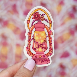Puede incluir: Una pegatina de linterna rosa y naranja con un diseño floral y estrellas. La linterna tiene un asa en forma de corazón y está decorada con enredaderas y hojas.