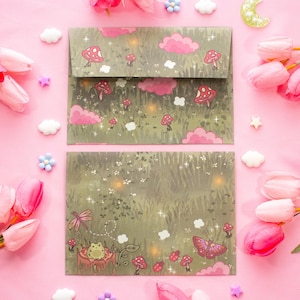 May include: Two stationery envelopes with a whimsical forest design featuring mushrooms, clouds, and a frog. The envelopes are set against a pink background with pink tulips and decorative accents.