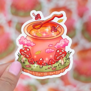 May include: A pink and orange cauldron with a wooden spoon and a golden liquid inside. The cauldron is surrounded by pink clouds and mushrooms with white spots. The background is a blurry image of pink and orange stickers.