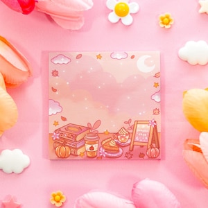May include: A square, pink notepad with a fall-themed illustration featuring a pumpkin cafe, books, and a coffee cup. The notepad is surrounded by pink flowers and decorative elements, creating a whimsical aesthetic.
