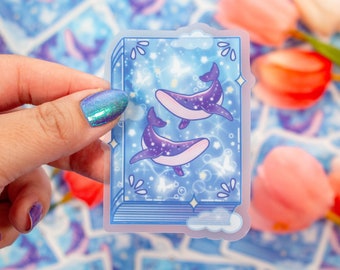 May include: A blue and purple whale sticker with a sparkly background. The sticker is shaped like an open book with a cloud on top. The sticker is on a white background.