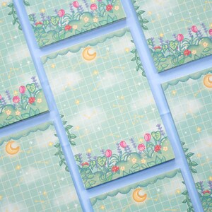 K&ouml;nnte beinhalten: Ein Set aus sechs pastellblauen Notizbl&ouml;cken mit einem Rastermuster und einem floralen Rand. Jeder Notizblock zeigt einen Halbmond und Sterne.