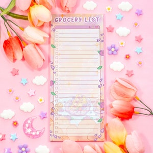 May include: A pastel-colored grocery list notepad with the words "Grocery List" at the top. The notepad is decorated with floral and cartoon elements. The background is pink, with decorative flowers, stars, clouds, and a crescent moon.