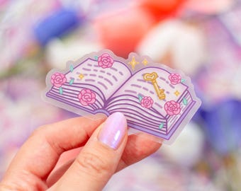 Può includere: Un adesivo trasparente con un libro viola con fiori rosa e una chiave dorata. Il libro è aperto e le pagine sono visibili. L'adesivo è su una mano.