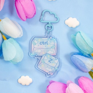 May include: A light blue acrylic keychain featuring two connected envelope-shaped charms with a pastel cloud and star pattern. The top envelope displays the text 'ctrl' and the bottom envelope displays the letter 'z'. It has a cloud-shaped clasp.