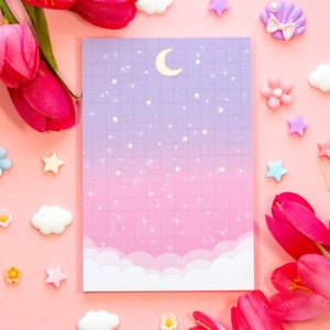 May include: A pink and purple notepad with a grid pattern and a crescent moon and stars design. The notepad is surrounded by pink flowers, clouds, and stars.