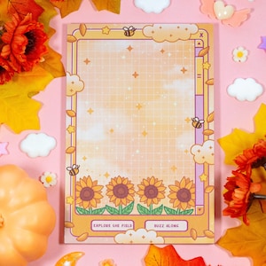 May include: A yellow and orange notepad with a grid pattern and a cartoon bee design. The notepad has the text "Explore the field" and "Buzz along" on the bottom. The notepad is surrounded by fall leaves and other decorative items.