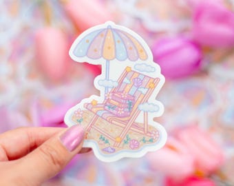 May include: A sticker featuring a beach scene with a striped lounge chair, books, and a colorful umbrella. The sticker has a pastel color palette with clouds, flowers, and seashells. The overall theme is summer and relaxation.