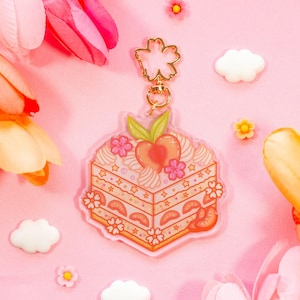 May include: An acrylic keychain featuring a square slice of cake decorated with pink cherry blossoms, whipped cream swirls, and a peach slice on top. The charm is attached to a gold-colored metal clasp shaped like a five-petaled cherry blossom.