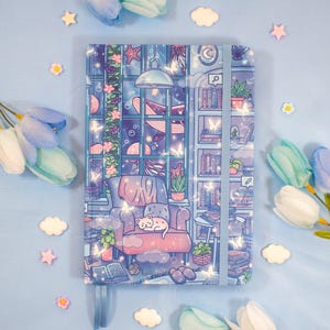 May include: A rectangular notebook with a blue elastic closure featuring a colorful illustration of a cozy room with a cat sleeping on a chair, bookshelves, a large window with a whale outside, and scattered small white butterfly and star accents.
