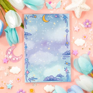 Puede incluir: Un cuaderno azul y morado con un diseño de cielo nocturno con estrellas, nubes y una luna creciente. El cuaderno tiene un borde blanco con un diseño caprichoso de estrellas fugaces.