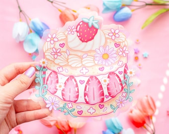 Può includere: Una torta rosa e bianca con una fragola in cima, circondata da fiori e viti. La torta è decorata con cuori e brillantini.