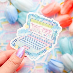 May include: A pastel-colored laptop sticker with a heart and design elements on the screen. The sticker has a light blue border with cloud and star accents. The keyboard is white, and the overall design is whimsical and cute.