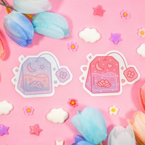 May include: Two pastel pink and blue tea bag stickers with a crescent moon, stars, and a flower design. The stickers are on a pink background with pastel blue and pink flowers, clouds, and stars.