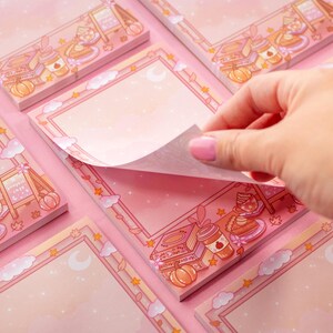 May include: Pink memo pads with a decorative border featuring illustrations of food, pumpkins, and stars. A hand is lifting a page from one of the pads. The pads have a blank space for notes.
