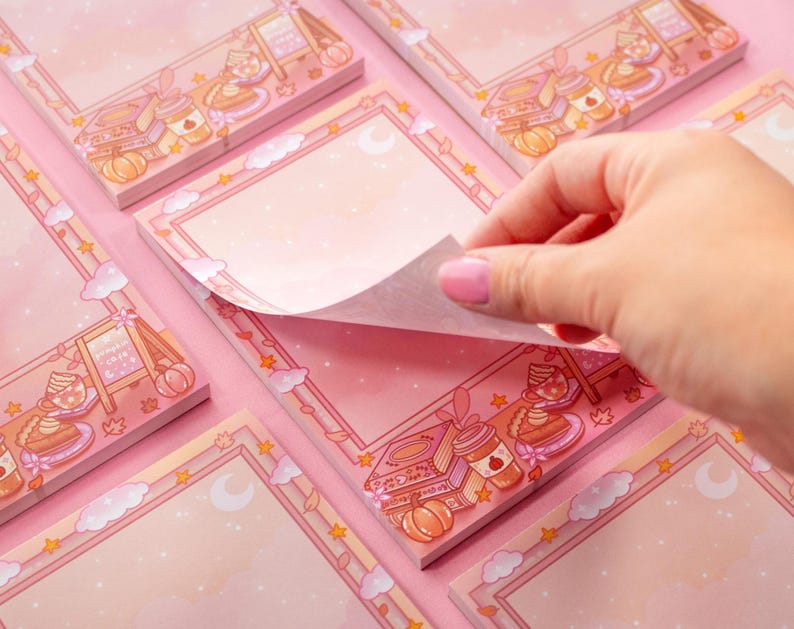 May include: Pink memo pads with a decorative border featuring illustrations of food, pumpkins, and stars. A hand is lifting a page from one of the pads. The pads have a blank space for notes.