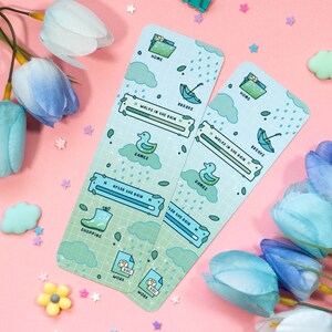 May include: Two blue and green illustrated bookmarks with a rainy day theme. The bookmarks feature a duck, an umbrella, a rain cloud, and a boot. The text on the bookmarks includes "Home", "Books", "Games", "Shopping", "Work", and "After the Rain".