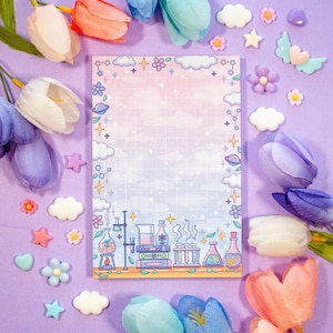 May include: A notepad with a science-themed illustration featuring beakers and flasks, set against a gradient background of pink, blue, and white. The notepad is surrounded by decorative flowers and star-shaped embellishments, all on a purple surface.