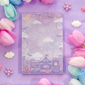 May include: A rectangular notepad with a purple background and a Halloween-themed illustration. The illustration includes a carnival scene, ghosts, bats, and a crescent moon. The notepad is surrounded by pink and blue tulips and decorative embellishments.