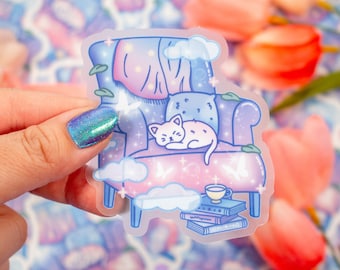 Puede incluir: Una pegatina transparente con un gato de dibujos animados durmiendo en un sillón morado con un degradado rosa y azul. El sillón está decorado con nubes, estrellas y mariposas. Una taza de té se encuentra sobre una pila de libros.