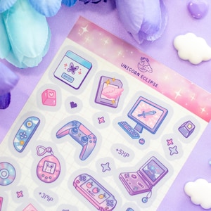May include: A sheet of pastel pink and purple holographic stickers featuring a variety of gaming-themed illustrations, including a game controller, a computer monitor, a Tamagotchi, and a handheld game console. The stickers are decorated with stars and hearts. The text "Unicorn Eclipse" is printed at the top of the sheet.