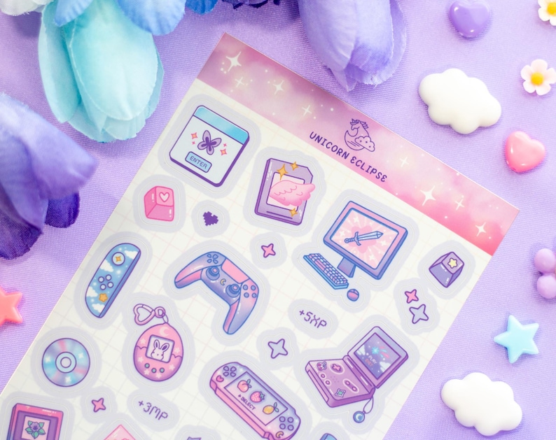 May include: A sheet of pastel pink and purple holographic stickers featuring a variety of gaming-themed illustrations, including a game controller, a computer monitor, a Tamagotchi, and a handheld game console. The stickers are decorated with stars and hearts. The text "Unicorn Eclipse" is printed at the top of the sheet.