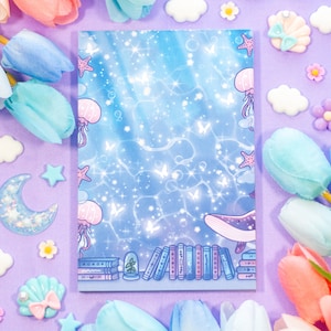 May include: A blue and white notebook with a whimsical underwater scene featuring a whale, jellyfish, and stars. The notebook is surrounded by pastel-colored flowers and other decorative elements.