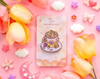 May include: A pink and gold enamel pin featuring a cup of coffee with whipped cream and a chocolate square. The pin is on a card with the text "Unicorn Eclipse" and surrounded by decorative flowers, stars, and clouds.