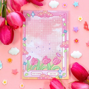 Puede incluir: Un bloc de notas rosa y blanco con un patrón de cuadrícula y un diseño de dibujos animados de nubes, flores y corazones. El bloc de notas tiene el texto "Pick Flowers" y "Let It Be" en él.