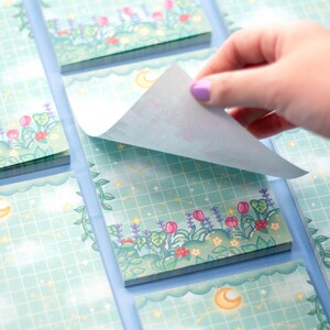 K&ouml;nnte beinhalten: Ein Stapel pastellblauer Haftnotizen mit einem verspielten Design mit Mond, Sternen und einem floralen Rand. Die Notizen sind mit einem Gittermuster verziert und haben eine hellblaue R&uuml;ckseite.