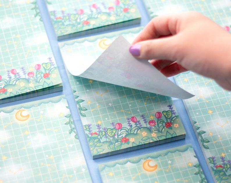 K&ouml;nnte beinhalten: Ein Stapel pastellblauer Haftnotizen mit einem verspielten Design mit Mond, Sternen und einem floralen Rand. Die Notizen sind mit einem Gittermuster verziert und haben eine hellblaue R&uuml;ckseite.