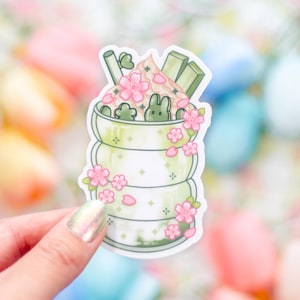 May include: A sticker featuring a matcha latte design with a bunny and cherry blossom accents. The sticker has a layered cup design with green and white colors, topped with pink whipped cream, green matcha sticks, and pink flowers.
