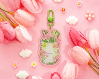 May include: A green and white acrylic keychain featuring a layered dessert design with a bunny and cherry blossoms. The keychain has a green clasp and is surrounded by pink tulips and decorative flowers on a pink background.