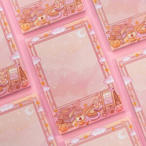 May include: A stack of pink notepads with a fall-themed border. The border features illustrations of pumpkins, coffee cups, books, and pie slices. The background has a soft, pink gradient with clouds, stars, and moons.