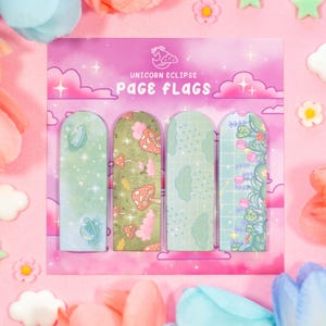 Può includere: Un set di quattro segnalibri decorativi con diversi disegni. I segnalibri presentano un pianeta, funghi, nuvole e fiori. Il testo "UNICORN ECLIPSE PAGE FLAGS" è stampato sulla carta rosa. I segnalibri sono circondati da fiori e stelle decorative.