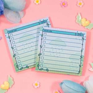 May include: Two square notepads with a blue and green design, resembling a computer window, are displayed. The pads feature a "TO DO LIST" header, checkboxes, and a rain and cloud illustration. The background is pink, with flower and heart decorations.