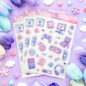 May include: A set of three clear sticker sheets with a pastel pink and purple background. The stickers feature a variety of cute cartoon illustrations of gaming consoles, controllers, and other gaming-related items. The text "Unicorn Eclipse" is printed on the top sheet.